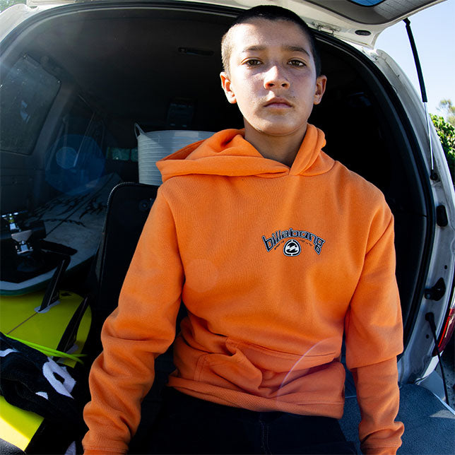 Boy wearing an orange hoodie with a logo, sitting inside an open car trunk.