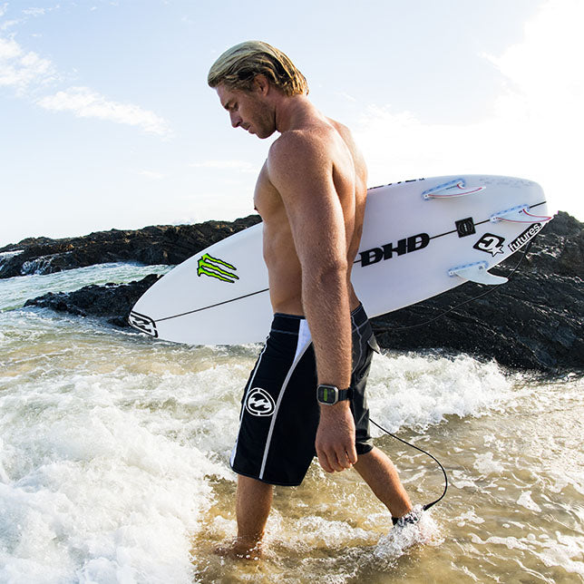 Man walking out of the ocean with a surfboard, wearing black board shorts with a logo.