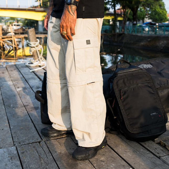 A man standing on a wooden dock wearing beige zip off cargo pants with black shoes, next to a black backpack, in an outdoor waterfront setting.