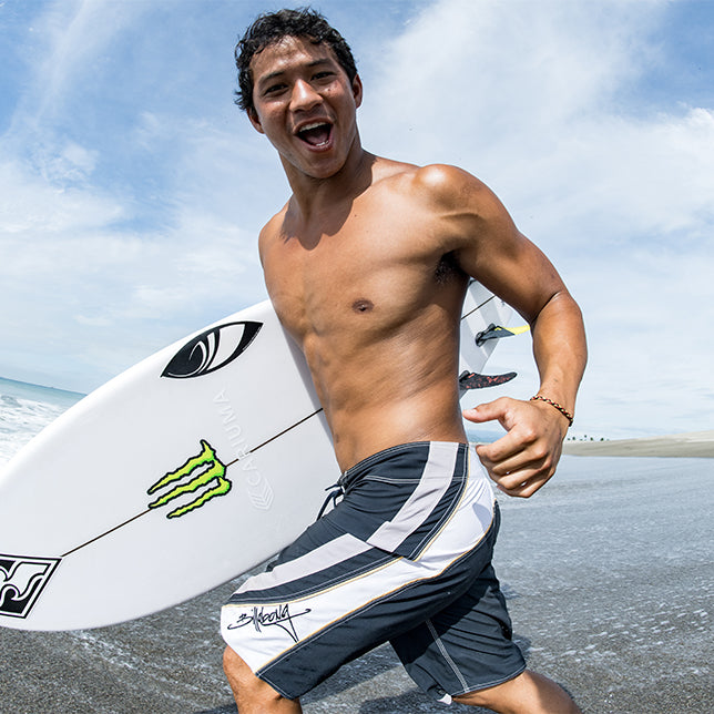 A man in board shorts holds a surfboard and smiles while walking on a sandy beach near the ocean, under a partly cloudy sky.