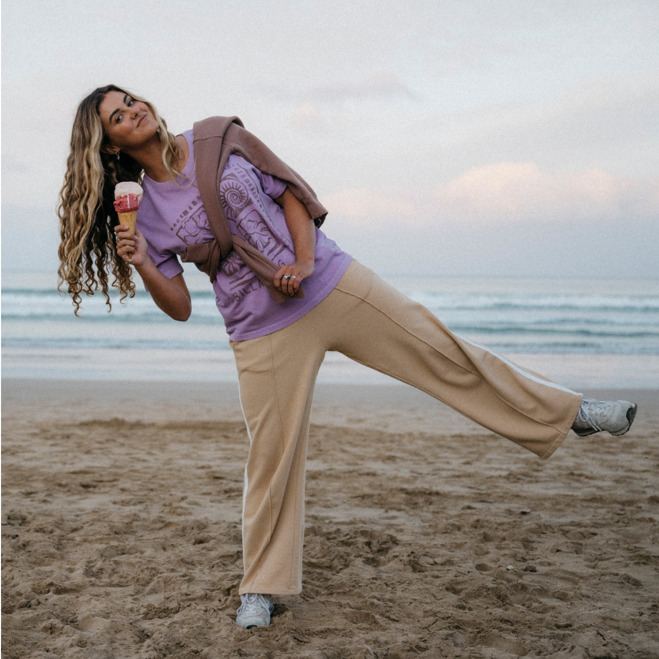 Woman on a beach wearing beige wide-leg track pants, a purple graphic T-shirt, and white sneakers, holding an ice cream cone.