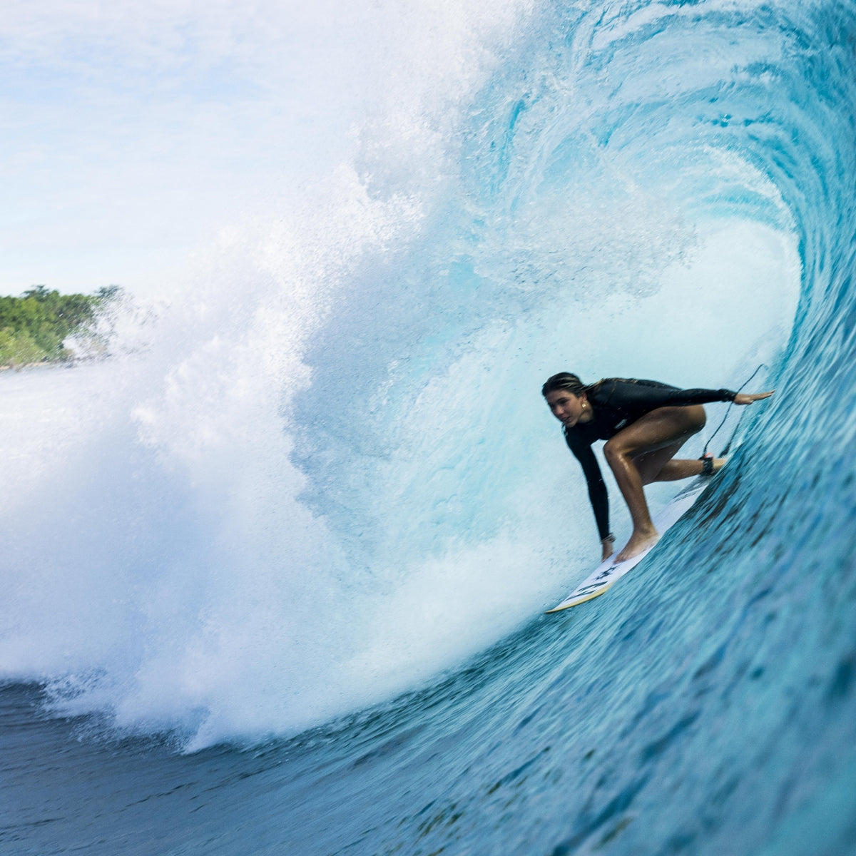 Action shot of a female surfer inside a barrel wave wearing a sleek black, long-sleeve women's springsuit wetsuit while carving on a white surfboard.