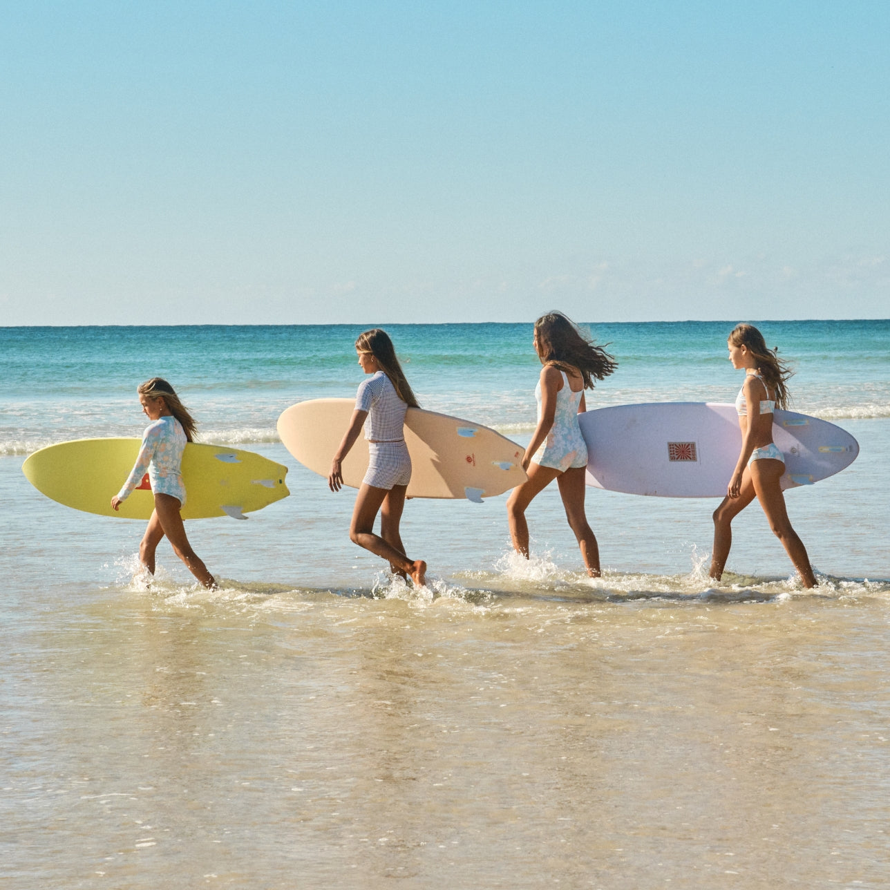 Four girls walking through shallow ocean water carrying colourful surfboards on a sunny beach.